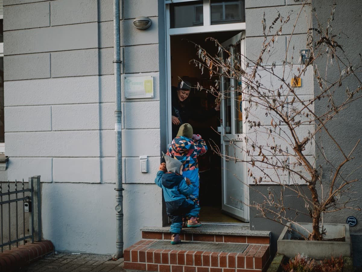 Kinder werden von einer Erzieherin am Eingang des Kinderladens begrüßt